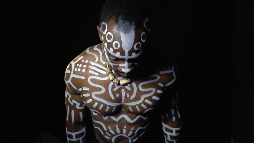 A black man in white patterns on his body stands with his eyes closed and exhales heavily, slow motion