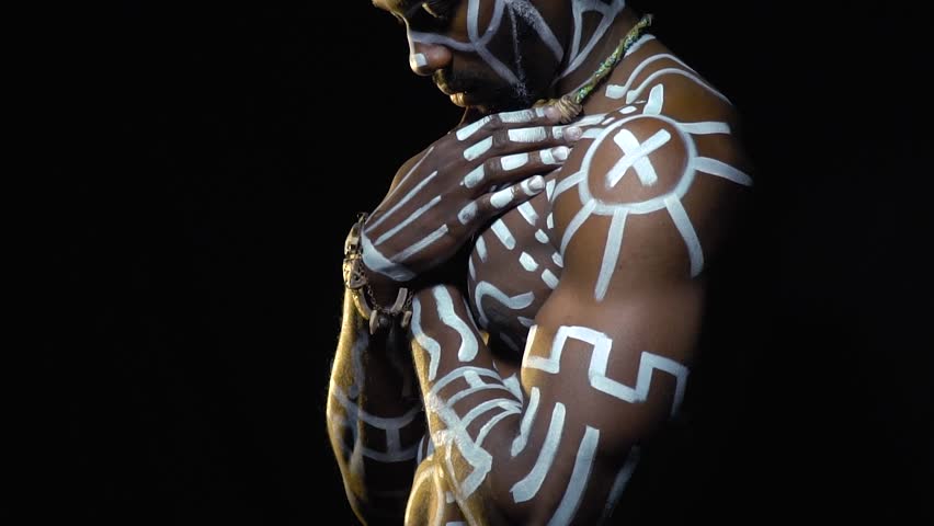 A serious black man in white paint patterns on his body stands in the dark