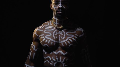 A black man in white patterns and a ritual hat praying on a black background, slow motion