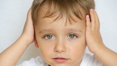 A close-up of a child looks thoughtfully at the camera, holding his hands behind his head, closing his ears. The child is thoughtful, does not want to hear anyone