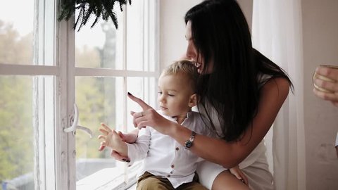Young mother is sitting with her son on the window sill decorated with Christmas wreath and looking outside. They are talking and smiling. Happy family at home