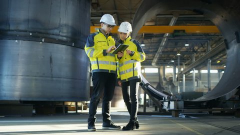 Male and Female Industrial Engineers in Hard Hats Discuss New Project while Using Tablet Computer. They Make Showing Gestures.They Work at the Heavy Industry Manufacturing Factory. Long Shot.