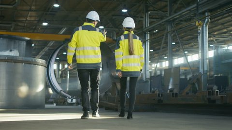 Back View Shot of Male and Female Industrial Engineers Having Discussion While Walking Through Heavy Industry Manufacturing Factory. Big Metalwork Constructions, Pipeline Elements Lying Around.