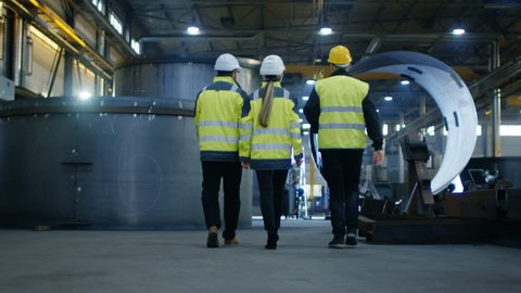 Following Shot of Three Engineers Walking Through Heavy Industry Manufacturing Factory. In the Background Welding Work in Progress, Various Metalwork, Pipeline/ Barrel Components. Slow Motion.