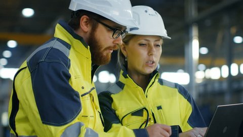 Portrait of Male and Female Industrial Engineers in Hard Hats Discuss New Project while Using Laptop. They Wear Safety Jackets.They Work at the Heavy Industry Manufacturing Factory. Long Shot. 4K.
