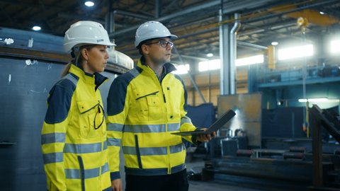 Male and Female Industrial Engineers in Hard Hats and Safety Jackets Discuss New Project while Using Laptop. They Walk Through on a Heavy Industry Manufacturing Factory with Metalwork Components.