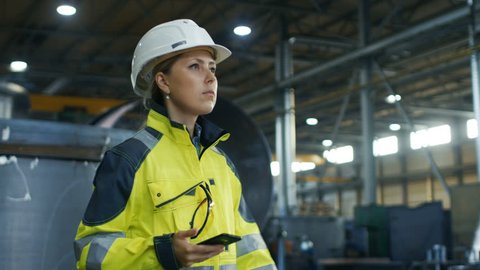 Female Industrial Worker in the Hard Hat Uses Mobile Phone While Walking Through Heavy Industry Manufacturing Factory. In the Background Various Metalwork Project Parts Lying. Shot on RED EPIC-W 8K 