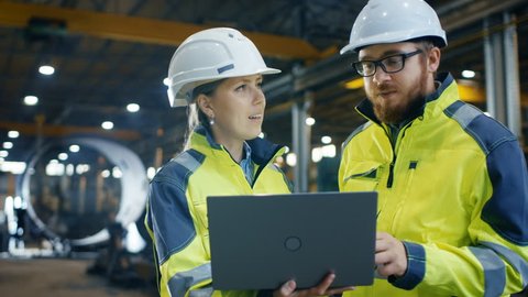 Inside the Heavy Industrial Factory Female Industrial Engineer Holds Laptop and Has Discussion with Project Manager. They Wear Hard Hats and Safety Jackets.  Shot on RED EPIC-W 8K Helium Cinema Camera