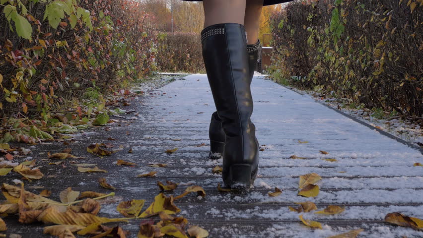Young slim beautiful woman is walking on snowy path with bushes in park at sunny winter day. Sexy legs close-up. Late autumn, snow. Slow motion. Steadicam low angle shot. Camera is following model.