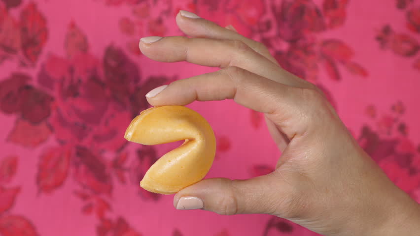Female hand holds up fortune cookie against pink floral background.
