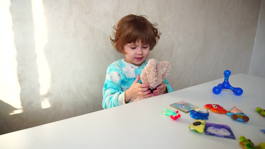 Little girl sitting holding plush rabbit in hands and adjusting decoration for bunny. Girl in blue pajamas with short haircut and curly hair sitting in bright room at white table. On table bright