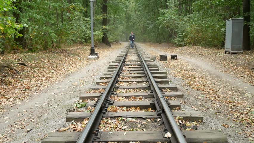 Young man walks between the railway rails. A railroad station in forest in the autumn. Student traveler with a black backpack and in blue jacket walks away from the camera