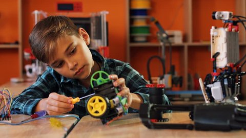 Portrait shot of nice blonde boy with the screwdriver assembling a robot and smiling straight into the camera in the modern comfortable playing space of classroom.