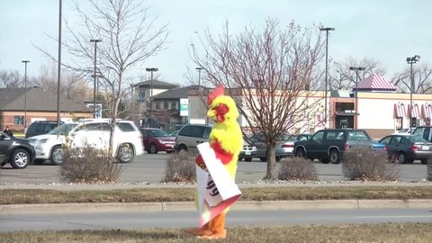 Man walks roadside in a yellow chicken suit selling restaurant specials with sign, getting attention of passing cars in sun.