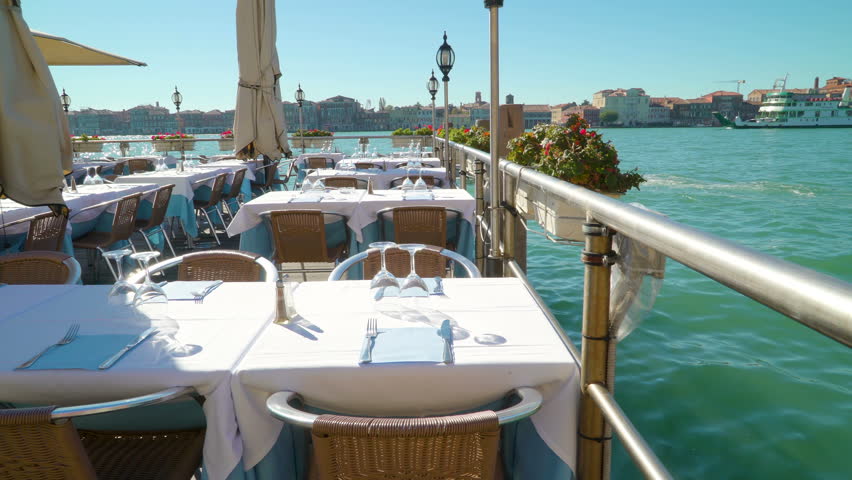 A view of the floating restaurant on the port in Venice with lots of tables with table napkins wine glass and spoon and fork and the ship on the background