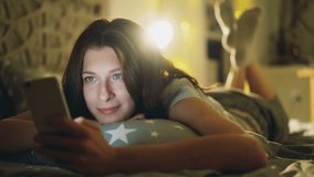 Attractive smiling woman using smartphone for sharing social media lying in bed at home at night