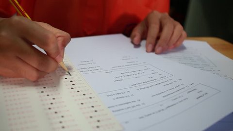 Students hand testing doing examination with pen drawing selected choice on answer sheets in school exams, blur pupils college background. Education system tests concept.Students hand testing exam