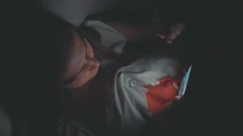 Woman using cellphone on bed at night. Young woman lying in bed and using smart phone before sleep at dark room, top view.