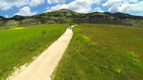 Top view of a plateau with greenery and a natural lake. Mountains in the background and view from the drone rising slowly. People walking on white gravel road