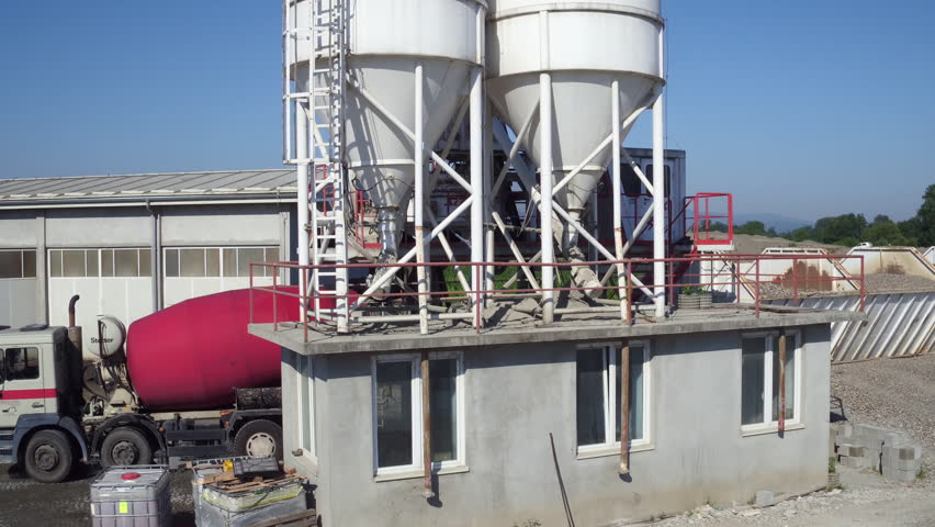 Mixer truck loading concrete from cistern, aerial view, camera motion over tank to countryside landscape with villages, mountain, forest and fields, bulldozer pushing sand on heap, building industry