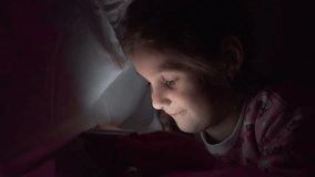 Small cute caucasian girl is looking at a smartphone cartoon lying on a bed under a blanket at night.