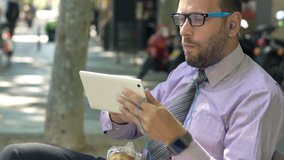 Businessman with sandwich watching movie on tablet sitting on bench in city