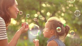 Happy Mother and her little Daughter playing together, blowing and catching soap bubbles.Beauty Mum and her Child in Park together. Family, Mother's Day Joy. Mom and Baby. Mother's Day celebrating. 4K