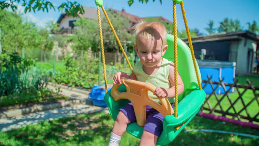 Tired little baby girl is rocking on a colourful plastic swing while a woman is working in a garden.