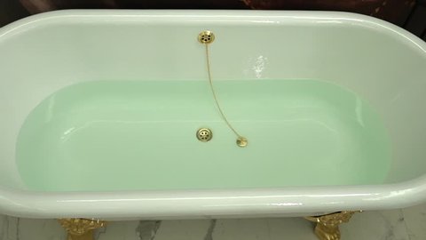 A vintage glazed ceramic iron clawfoot bathtub with golden-colored overflow opening with chain connected to tub drain stopper. A white bath on golden-colored claw feet filled with transparent water.
