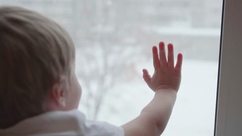 A child stands near a window and watching snow falling on the street. The closeup hand on the glass window