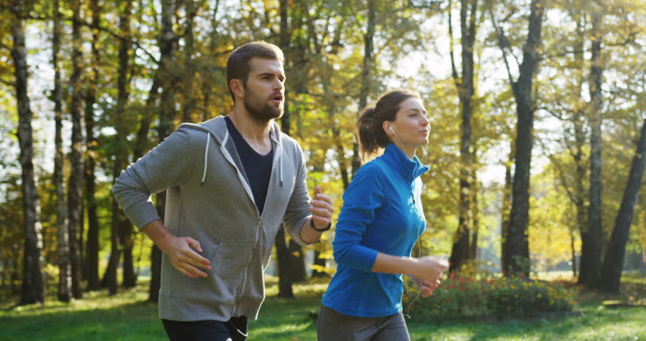 Slow motion from the side of young sporty couple jogging in the sunny park. The nice weather in early autumn. Outside