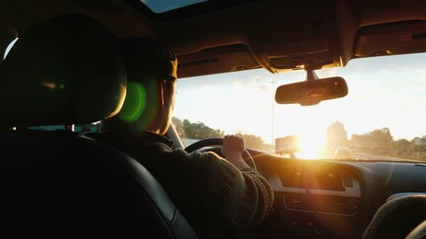An Asian man drives a car along the highway, the setting sun shines in the windshield. Back view