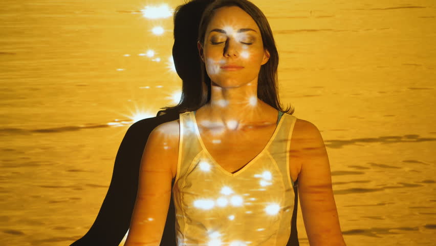 Attractive woman in yoga wear relaxes against wall with eyes closed. Peaceful, calm, meditation. Video of defocused sparkling and sunlit, golden slomo water projected onto model and background.
