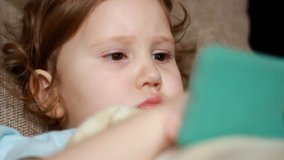Funny child looks at the phone screen and plays downloaded application on a smart phone close-up. A little cute girl lies in sofa in a living room, looking cartoon and playing the game. Close-up.