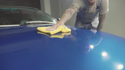 Master with tatooing hands finish polish the deep blue sport car and wiping the hood by cloth at a car workshop, slow motion