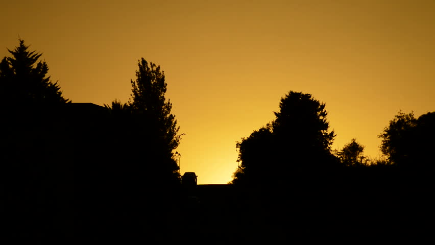 Cars coming down the hill with the sun hiding between houses and trees at sunset