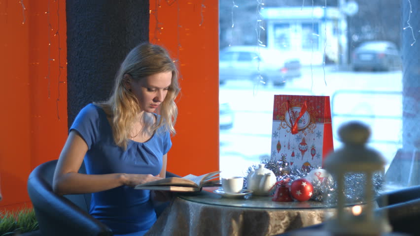 Girl with a book in a cafe. Slow motion. A young girl is sitting in a cafe and reading a book. 
On the table lay new year