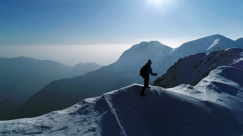 Aerial - Young man hiking on top of snowy mountain at beautiful winter sunset. Male mountaineer with trekking poles and a backpack walking on mountain ridge in Julian Alps