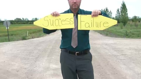 Businessman holds up pointed yellow signs at dirt road intersection reading success in one hand and failure in the other.