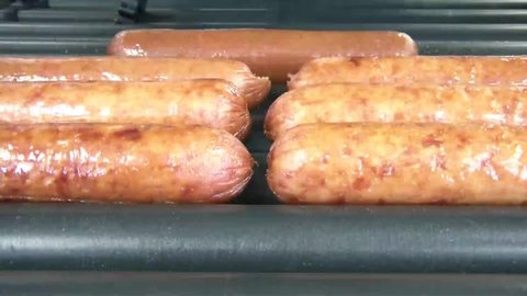 Brightly lit hot dogs and sausages rolling on heated metal with black plastic tongs picking one up during clip.