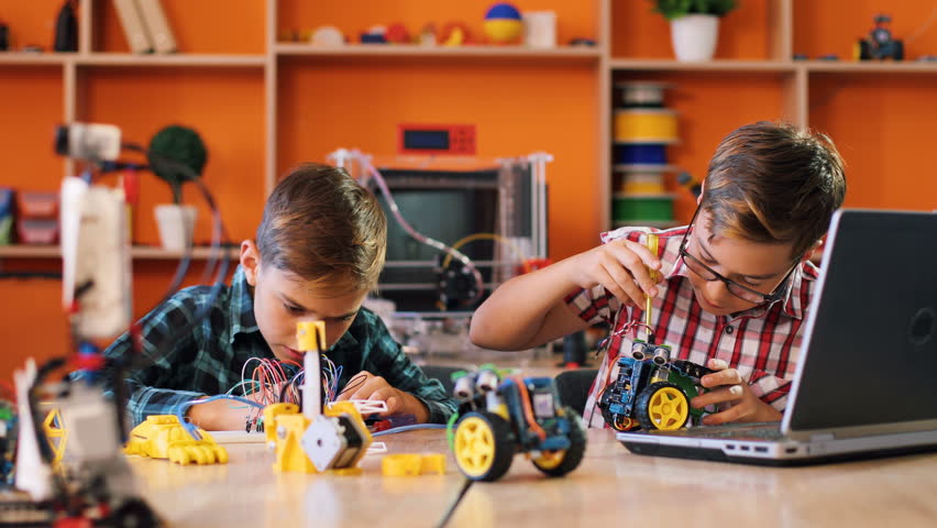 Two boys using screwdrivers to assemble robotic machines and watching how to do it on the laptop in the modern comfortable playing space.