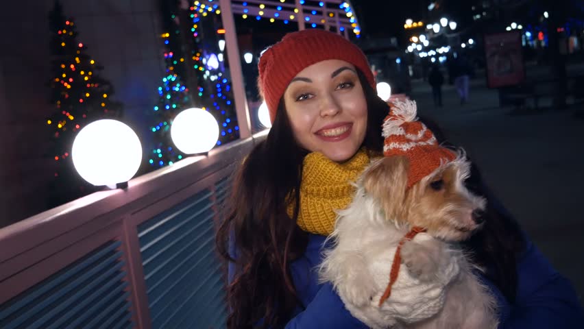 Young girl with a cute dog on the street. A beautiful girl is holding a dog in a hat. The girl smiles and dances.
Evening. The city is decorated with New Year