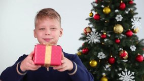 Happy caucasian smiling boy holds small red Christmas gift box in hands. Kid gives present to camera. Real time full hd video footage.