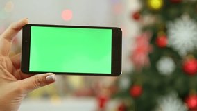 Closeup of female hands holding black modern smartphone in hands at glowing Christmas home interior. Adult woman uses digital device with blank green screen. Real time full hd video.