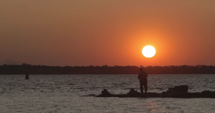 The amateur fisherman catches fish at sunset on a pier at the seashore. The man has a rest at the weekend at the lake in the open air, the fan of loneliness. Dawn background, card.