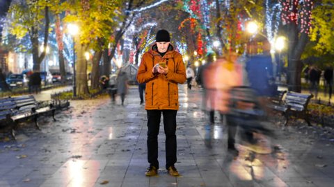 Timelapse of man standing still with smartphone on crowded walkway park boulevard with christmas garlands at night. People moving fast. Concept of flowing time and technologies