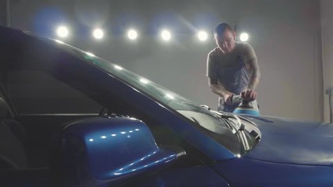 Master with tattooing hands polishes the deep blue sport car via polish mashine in a car workshop, slow motion