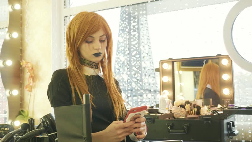 Young beautiful girl with halloween makeup sits on the chair and using smart phone at beauty salon.