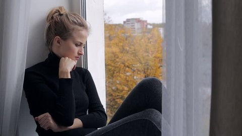 Young depressed and sad woman sitting on the windowsill