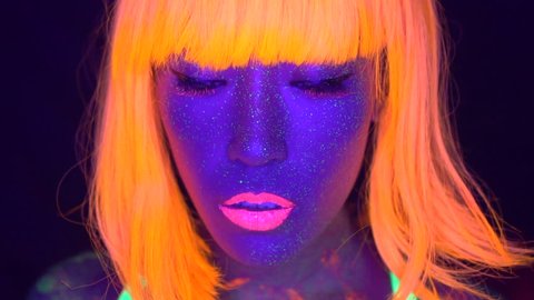 Closeup woman face with fluorescent make up in orange, wig, creative makeup look great for nightclubs. Halloween party, shows and music concept - slow motion video